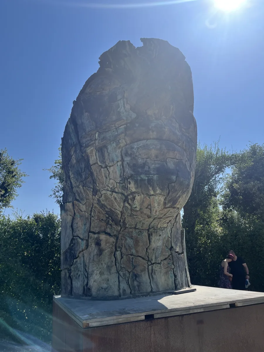 Tindaro Screpolato (1997) by Igor Mitoraj — the 407 cm bronze silhouetted against the sun in the Boboli Gardens, the cracked surface visible through backlit haze, a couple resting to the right for scale
