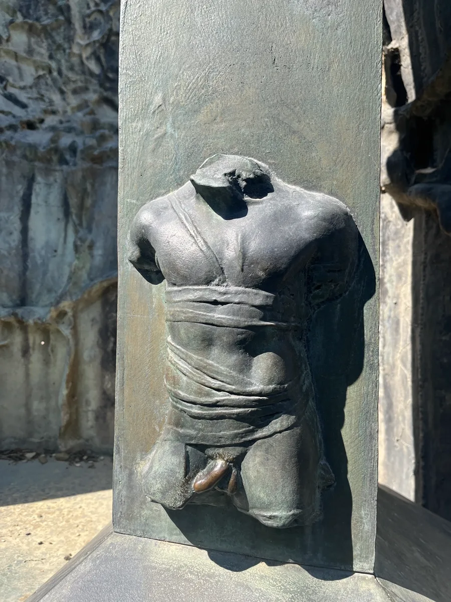 Detail of an Igor Mitoraj bronze torso figure set in an architectural niche, chest bandaged in cloth, framed by roughly textured bronze uprights — photographed during the same July 2026 Florence visit