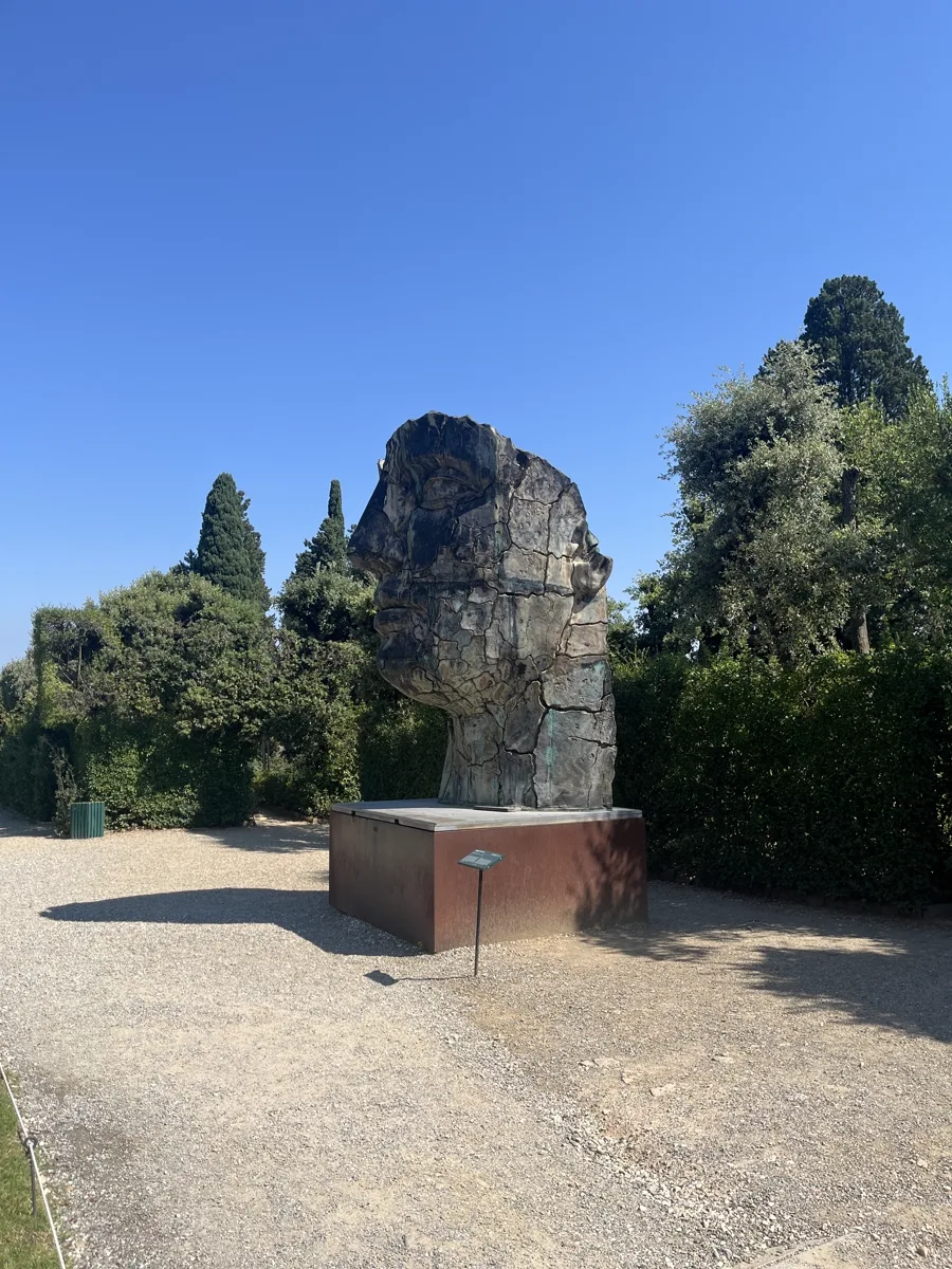 Tindaro Screpolato seen from the side across the Boboli path, its cracked profile rising above the hedge line under a clear sky, gravel walkway leading in from the left