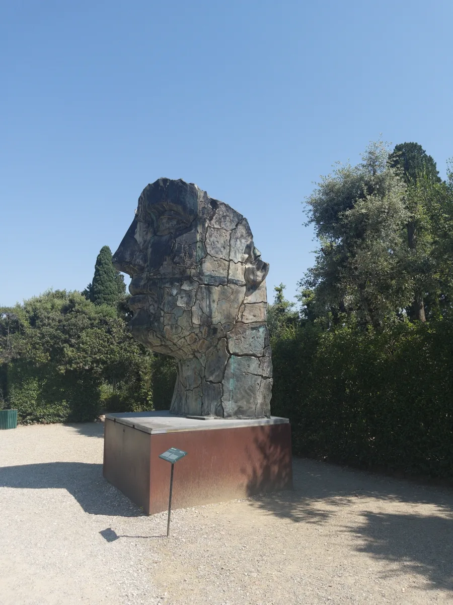 Ground-level view of Tindaro Screpolato at the far end of a Boboli walkway, cracked bronze catching side light against dark garden foliage